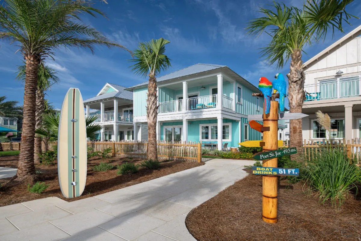 A colorful exterior view showcases a surfboard, a vibrant signpost, and playful parrots near a blue beach house, all surrounded by palm trees. The porch offers a cozy spot to watch the sunset, while the scene suggests easy access to both pool parties and beach adventures.