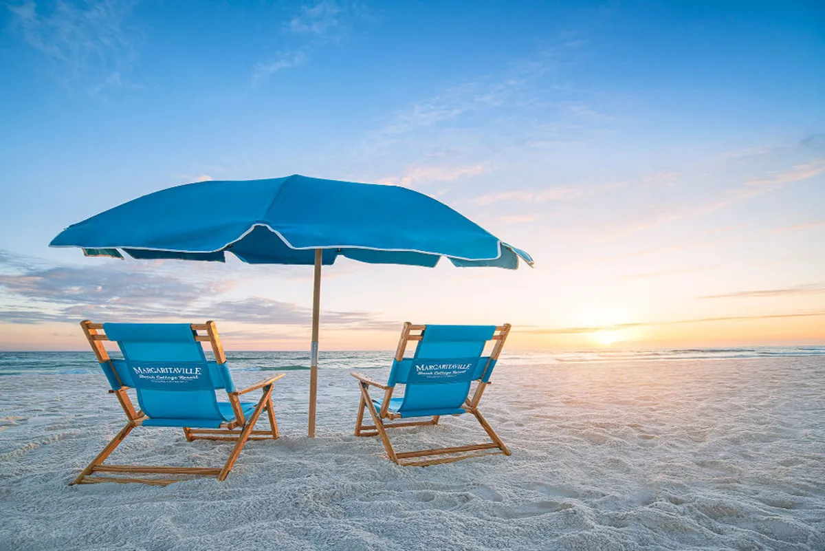 The hero image features two inviting blue Margaritaville-branded lounge chairs under a matching blue beach umbrella, set on pristine white sand facing the tranquil Gulf of Mexico. The scene captures a breathtaking sunset over the ocean horizon, suggesting peace, relaxation, and the quintessential beachfront experience.