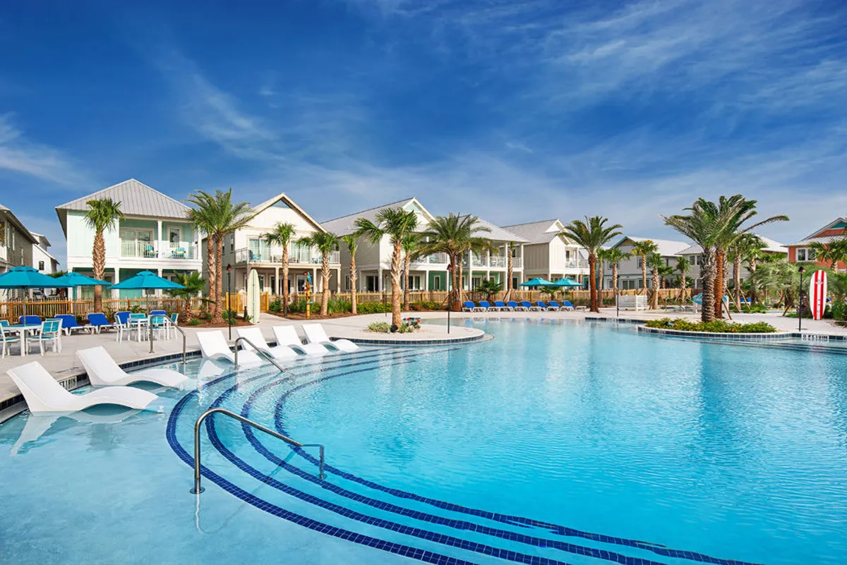 A large, modern resort-style pool is surrounded by rows of palm trees and two-story beachfront cottages with balconies. Colorful umbrellas and poolside seating invite guests to swim, sunbathe, and socialize, all set against the backdrop of the ocean and luxury accommodations.