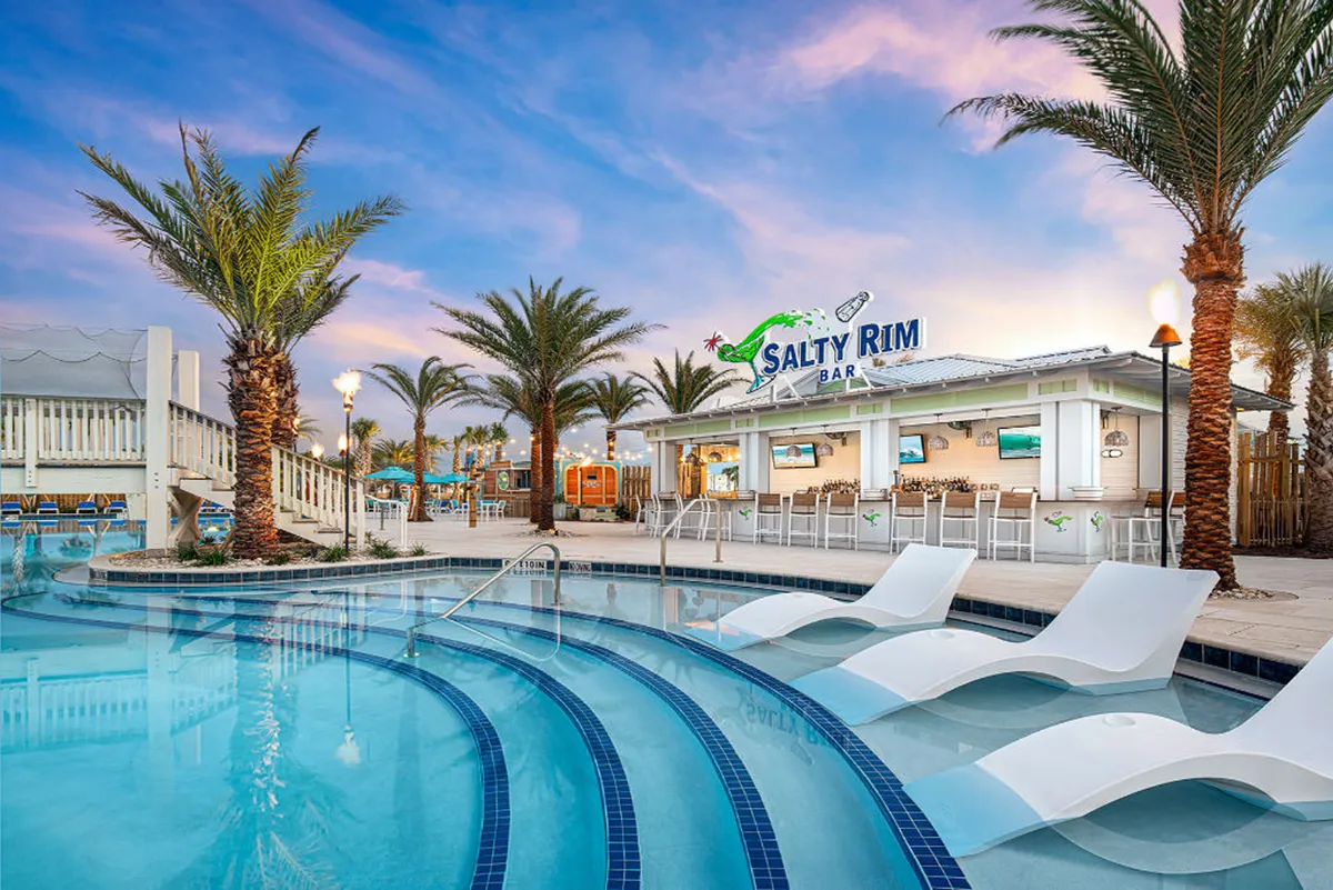 This image features the Salty Rim Bar & Grill poolside bar set against a heated resort pool with built-in lounge chairs. Palm trees frame the tropical setting, and wooden stairs lead to an elevated deck, hinting at more leisure spaces beyond. The scene is lively with guests enjoying poolside drinks and the inviting atmosphere of a full-service resort.
