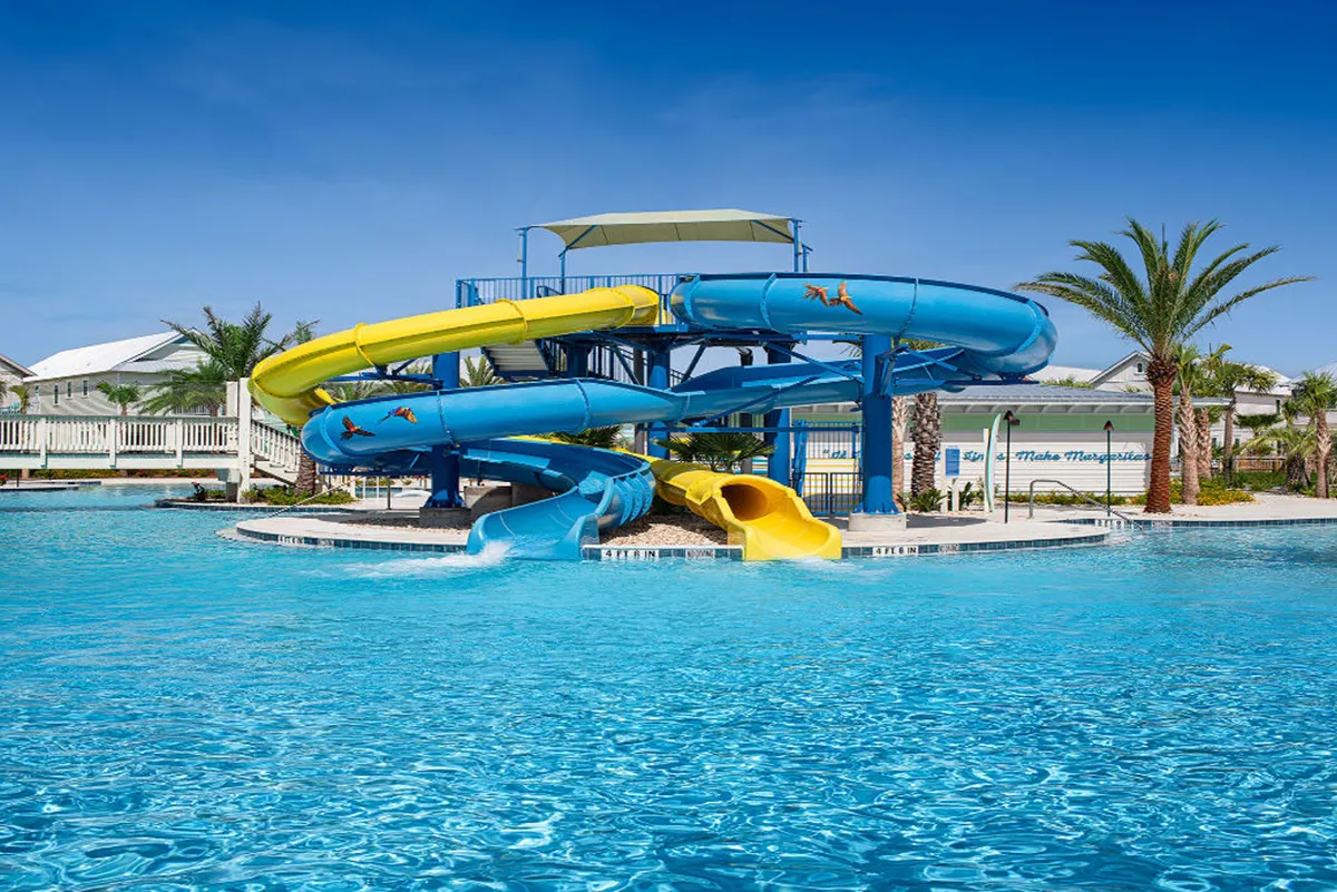 A striking view of large, colorful blue and yellow water slides emptying into a crystal-clear pool, surrounded by palm trees and sun loungers. The scene exudes excitement and is bustling with families and groups enjoying water activities under the sun.