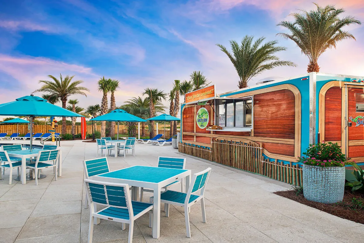 This lively scene shows turquoise and white outdoor tables and chairs arranged near a wood-paneled food and drink kiosk. Tall palm trees and bright blue umbrellas complete the tropical, social atmosphere, inviting guests to dine and relax outdoors in the resort’s vibrant communal spaces.