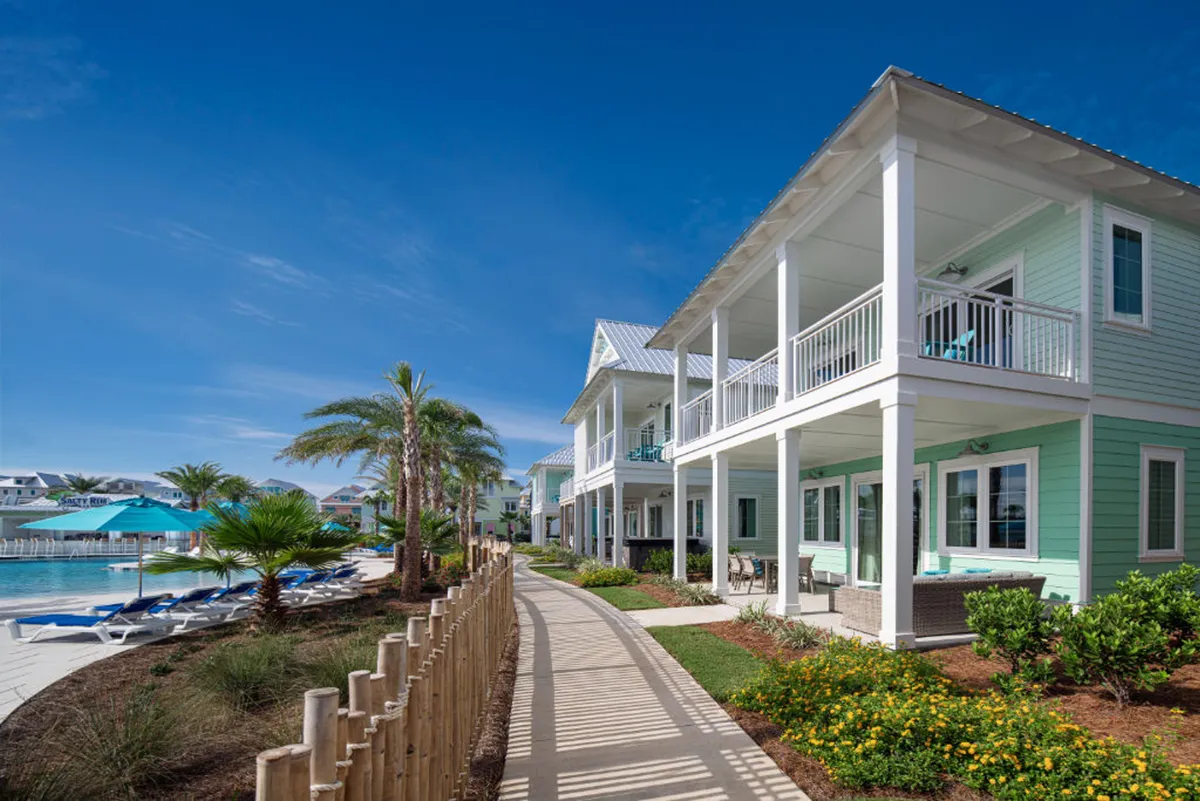 This image showcases a row of turquoise beach cottages with white trim and inviting balconies, with a well-kept pathway lined by natural wooden posts. The pool area with blue umbrellas and lounge chairs is visible, surrounded by palm trees and lush greenery. Guests can be seen relaxing or socializing on balconies and poolside seating, creating a sense of leisure and community.