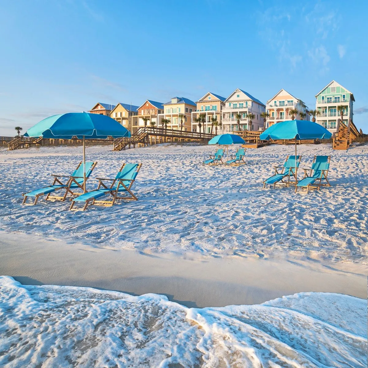 Rows of colorful beachfront houses with balconies and wooden staircases are set against a backdrop of a white sandy beach and gentle Gulf waves. Blue beach umbrellas and lounge chairs dot the shore, while several private wooden dune walkovers provide easy beach access. Guests are seen sunbathing, playing in the ocean, and socializing along the beach.