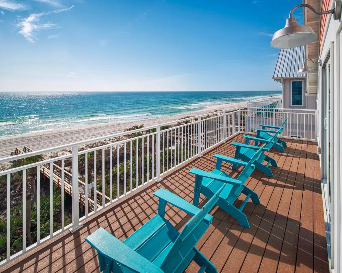 A private beachfront balcony featuring turquoise Adirondack chairs lined up to face an expansive view of the sandy beach and the Gulf of Mexico. The white railing and wooden deck create a fresh, coastal ambiance, perfect for enjoying the ocean breeze and morning sunlight.