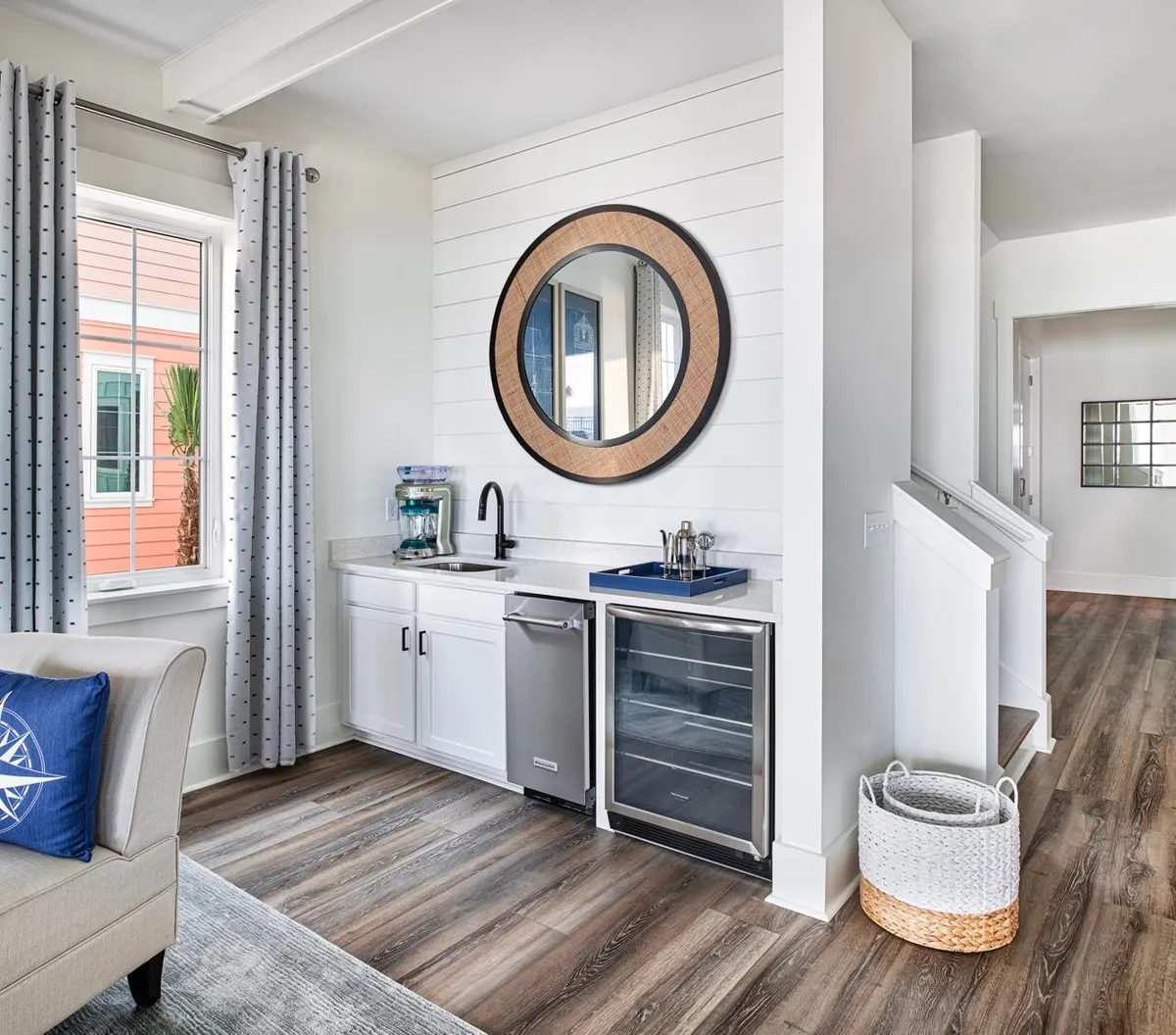 An inviting wet bar area with a sink and mini fridge, adorned with a round wicker-framed mirror and patterned curtains by a window, and an adjacent staircase leading to the upper floor. The space feels comfortable and casual, ideal for preparing drinks and gathering with friends or family.
