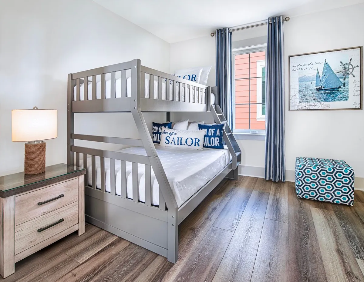 A cozy bedroom featuring a gray bunk bed with both twin and full size mattresses. Nautical-themed pillows add playful, beachy flair, while a framed sailboat artwork reinforces the coastal vibe. Natural light streams in from a window, highlighting the geometric ottoman and the tranquil, family-friendly ambiance.