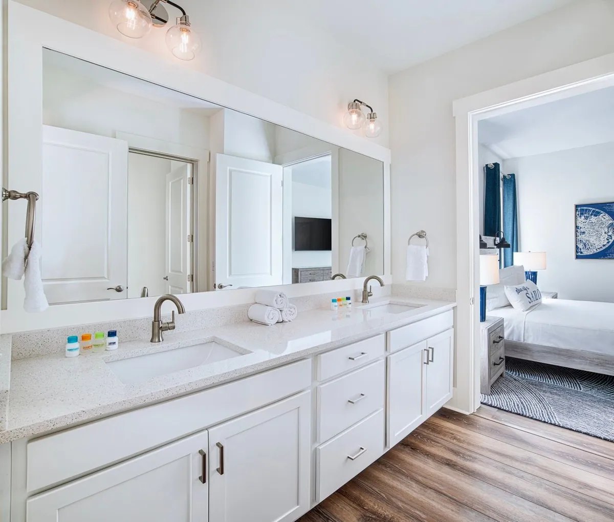 A bright, modern bathroom with a double vanity and sleek quartz countertop. Large mirrors reflect ample daylight, while rolled white towels and small toiletries add touches of spa-like luxury. An adjacent bedroom is visible through an open doorway, creating a sense of connection and openness.