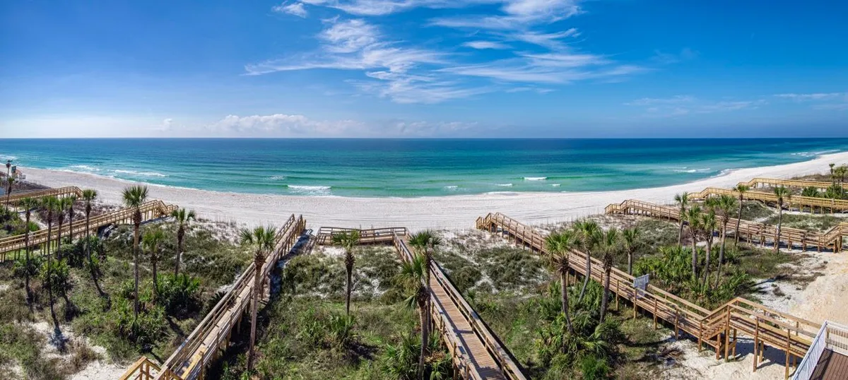A sunlit wooden boardwalk leads directly over the dunes to a vast stretch of white sandy beach and the turquoise waters of the Gulf of Mexico. The sky is clear with a few wispy clouds, and the untouched shoreline invites visitors for walks, swimming, or relaxation. The image embodies easy, private access to the beach and the open, welcoming feel of the coast.